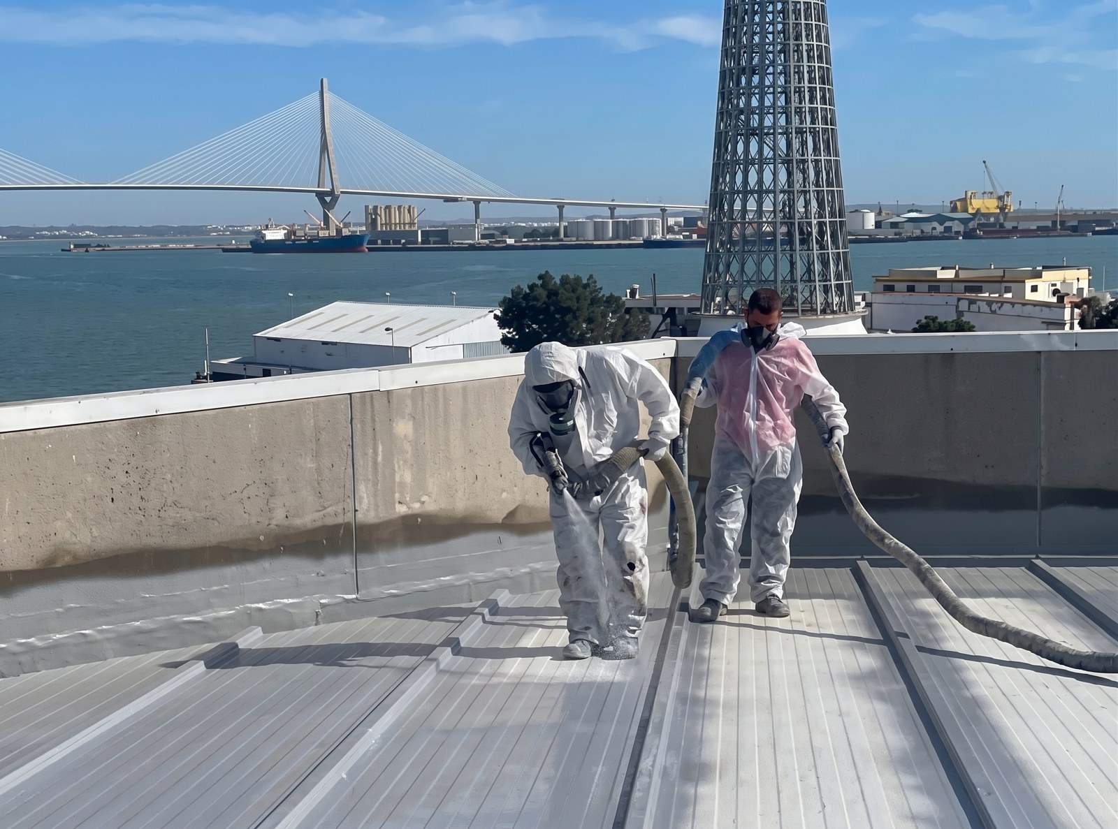 Dos operarios con equipo de protección proyectando poliurea en caliente sobre una cubierta metálica en Cádiz, con el puente de la Constitución de 1812 al fondo