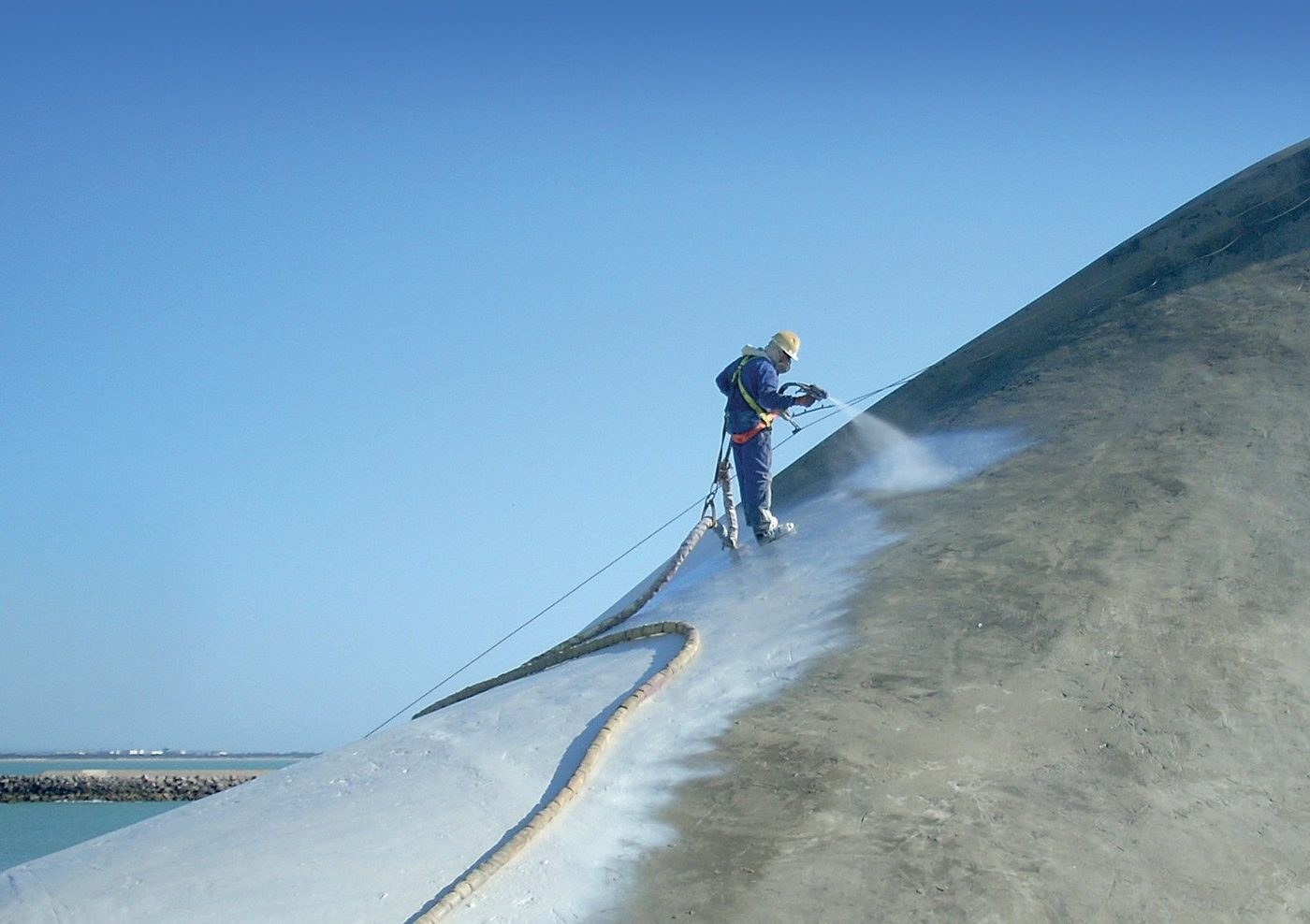 Operario asegurado proyectando poliurea sobre el talud de hormigón de un dique marítimo junto al mar